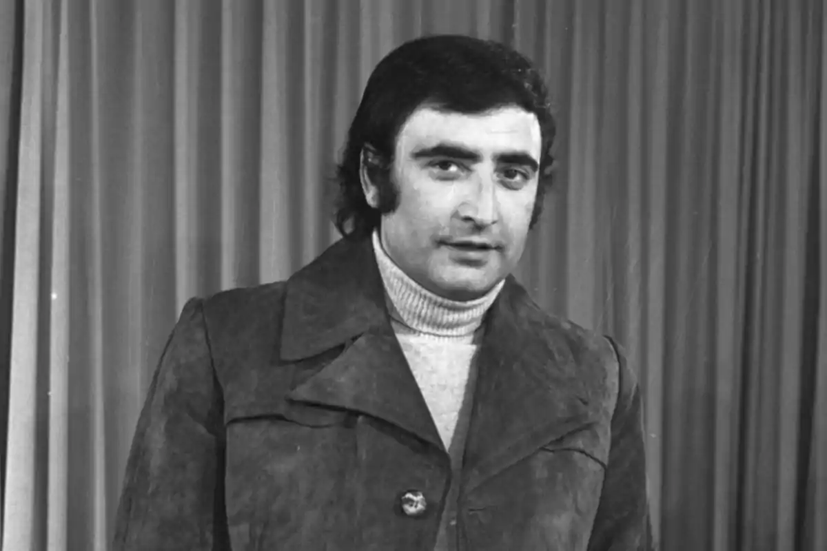 Hombre con chaqueta de gamuza y suéter de cuello alto posando frente a una cortina.