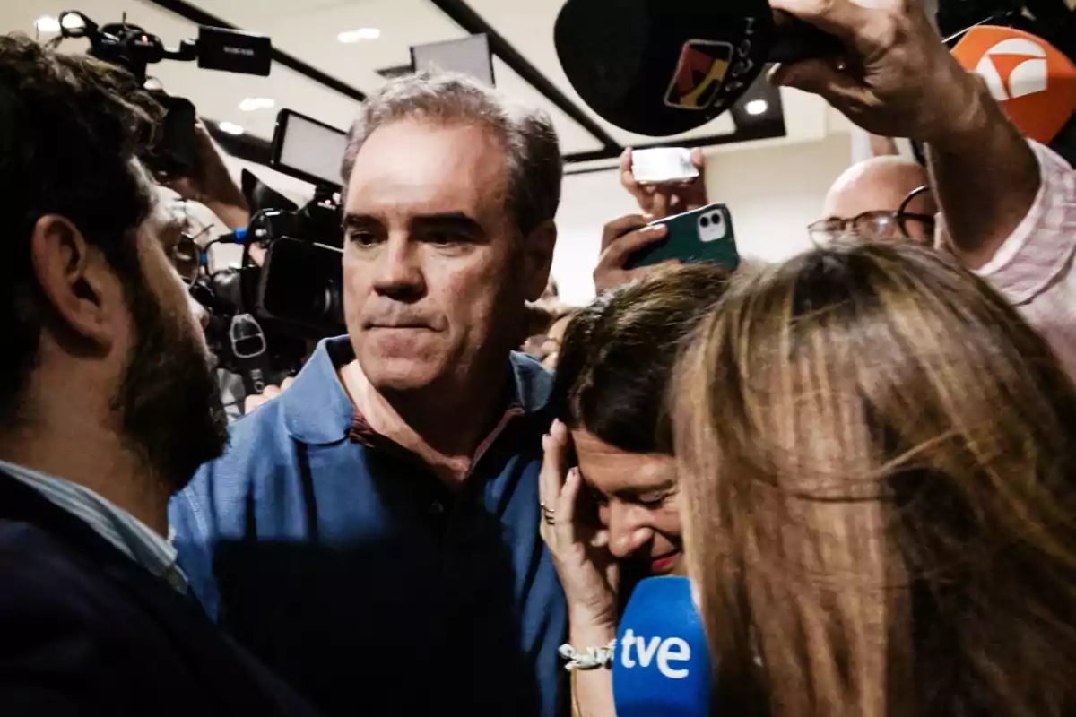 Un grupo de periodistas y camarógrafos rodea a una mujer que se cubre el rostro mientras un hombre la acompaña y varias cámaras y micrófonos apuntan hacia ellos