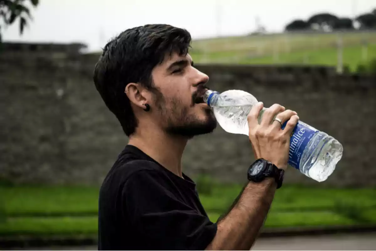 Un hombre con barba y cabello oscuro está bebiendo agua de una botella de plástico mientras está al aire libre.
