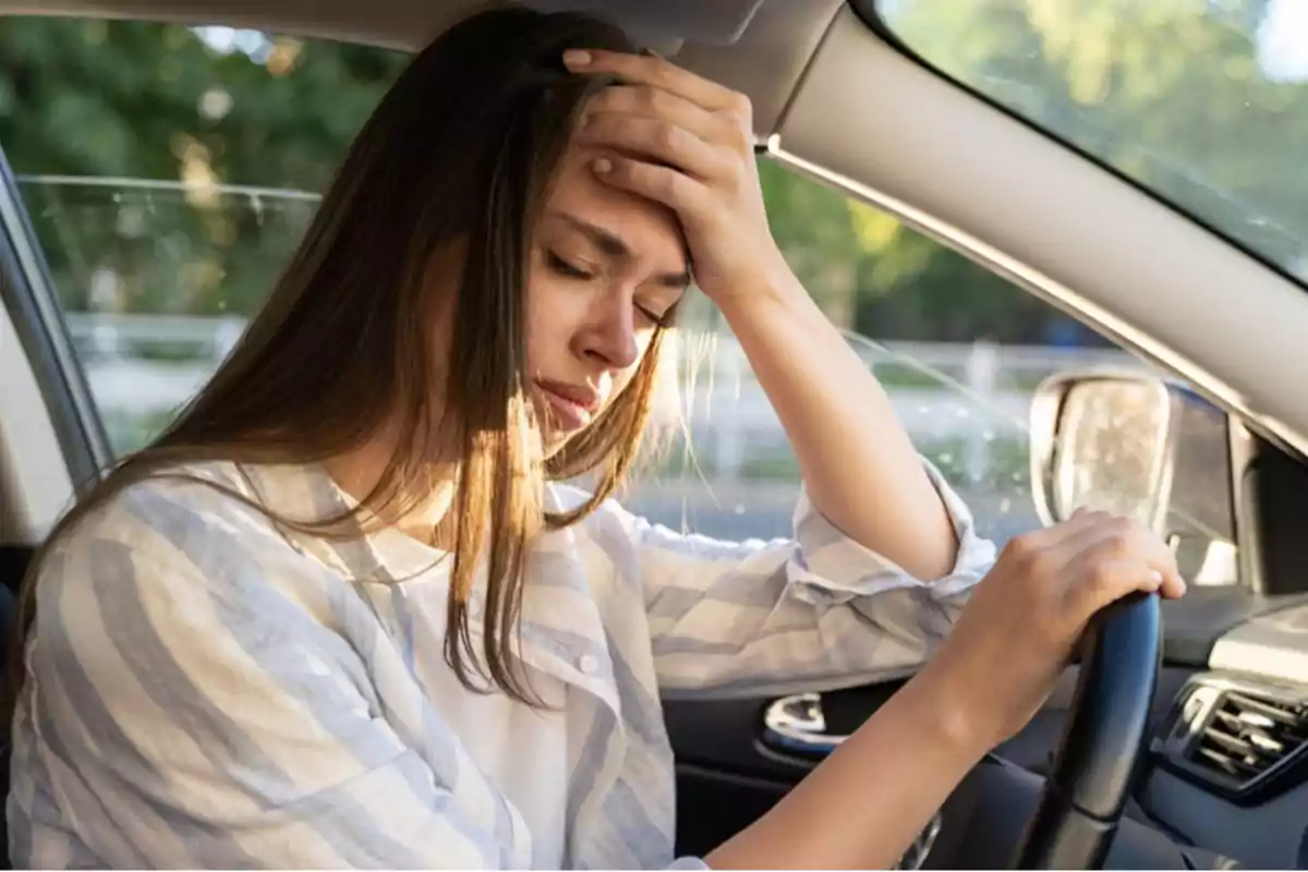 Mujer con expresión de cansancio o estrés sentada en el asiento del conductor de un automóvil, con la mano en la frente.