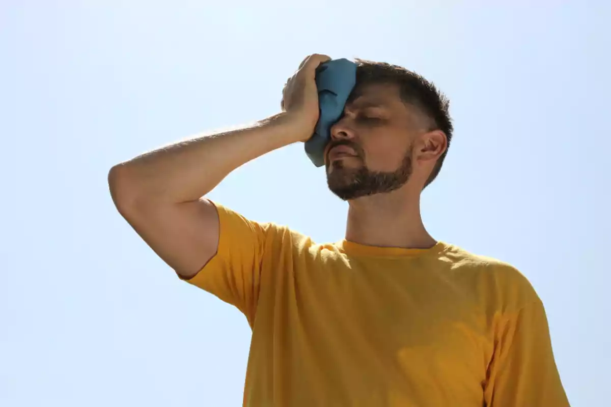 Hombre con camiseta amarilla aplicando una compresa fría en su frente bajo el sol.
