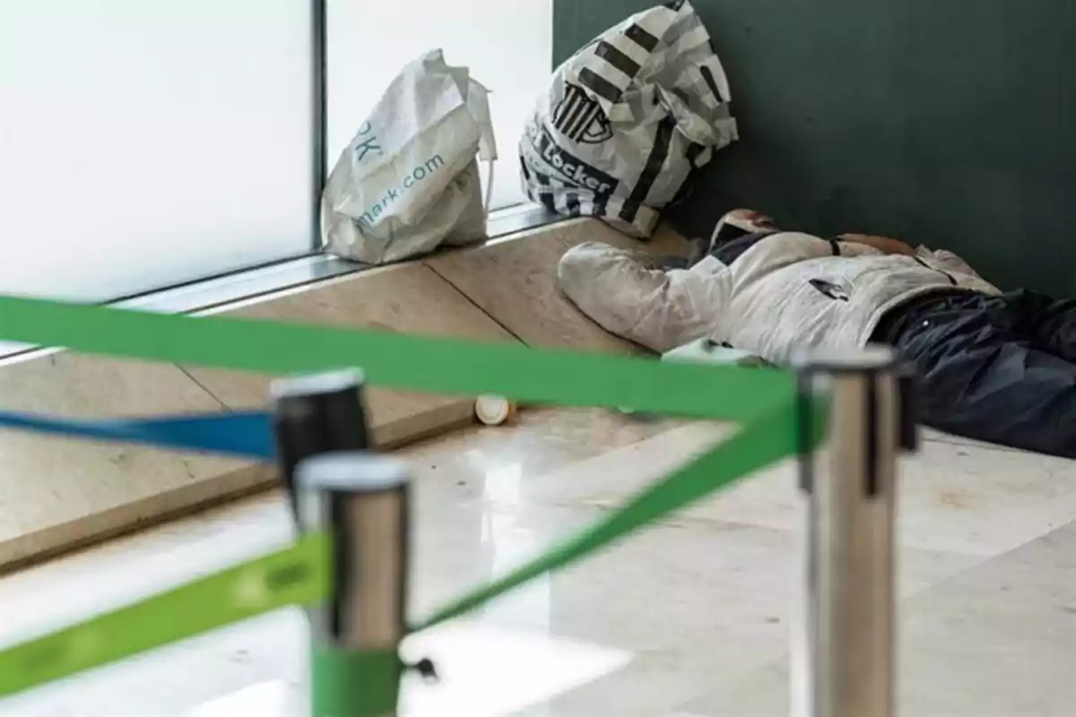 Persona durmiendo en el suelo junto a una pared con bolsas y cintas de seguridad en primer plano.