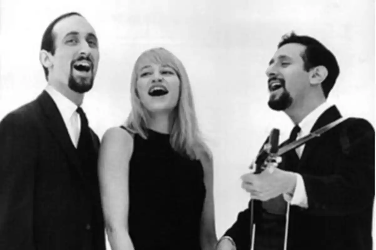 Tres personas cantando juntas, una mujer en el centro y dos hombres a los lados, uno de ellos tocando una guitarra.