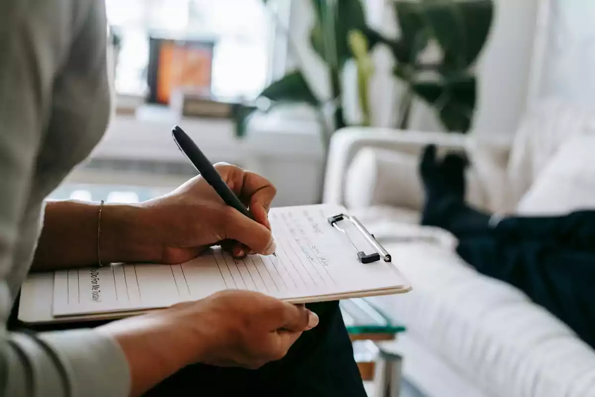 Persona escribiendo en una libreta con un bolígrafo mientras otra persona está recostada en un sofá.