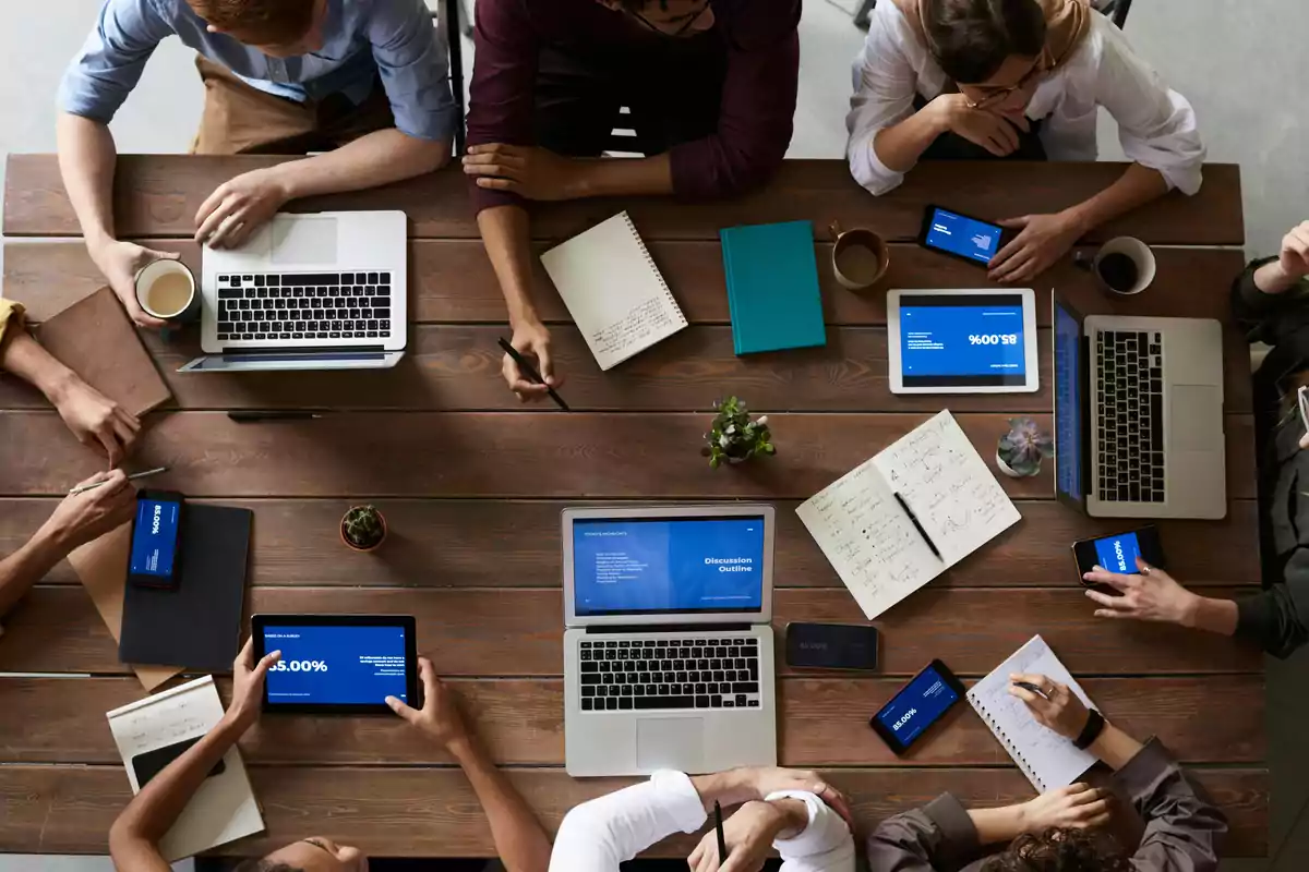 Personas reunidas alrededor de una mesa de madera trabajando con computadoras portátiles, tabletas, teléfonos y cuadernos
