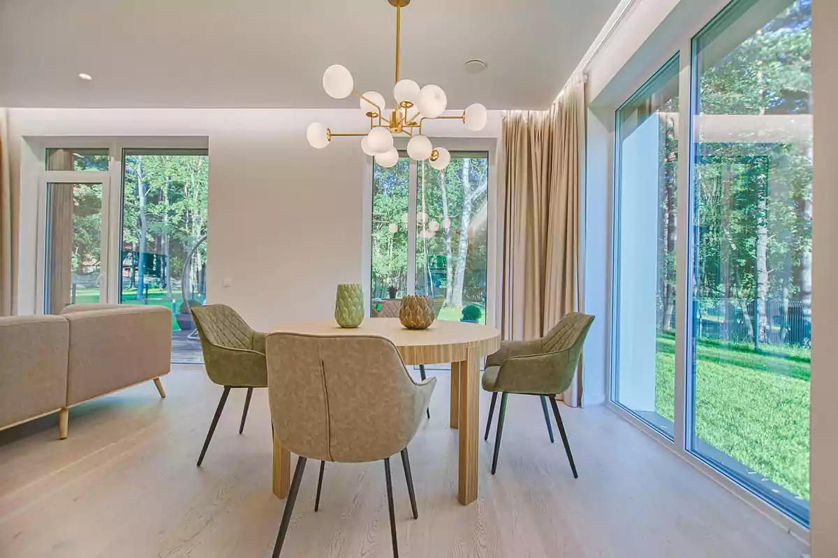 Comedor moderno con mesa redonda de madera, cuatro sillas tapizadas, lámpara de techo contemporánea y grandes ventanales que muestran un jardín exterior