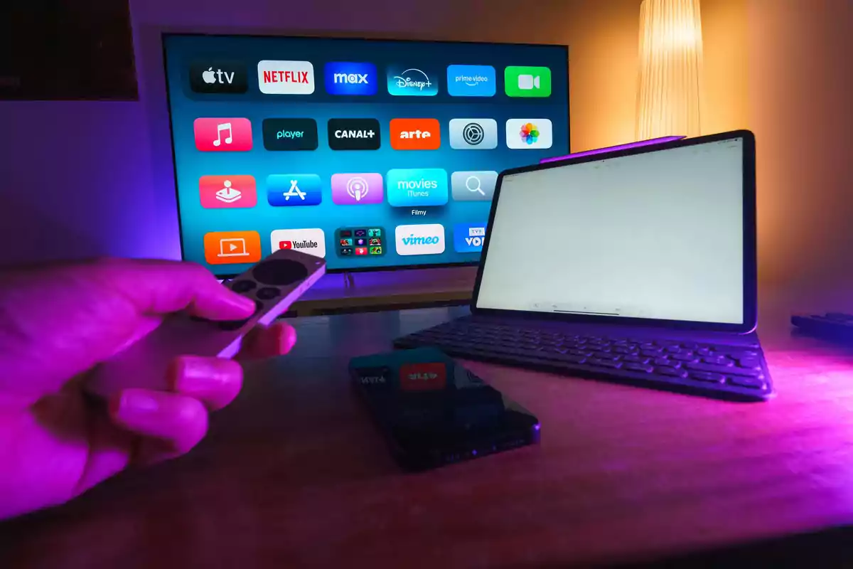 Persona usando un control remoto frente a una televisión inteligente con varias aplicaciones de streaming en la pantalla, junto a una tablet y un teléfono móvil sobre una mesa iluminada con luz morada.