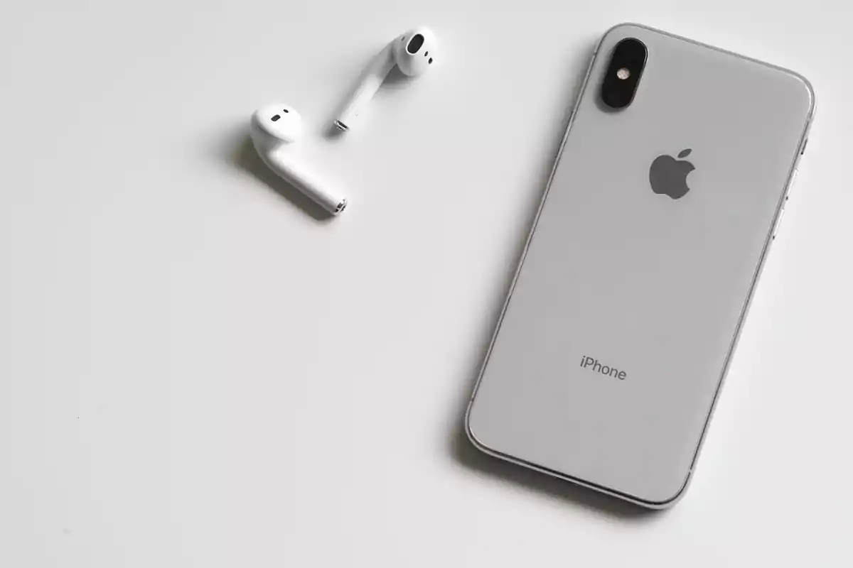 Un iPhone plateado junto a unos auriculares inalámbricos sobre una superficie blanca.