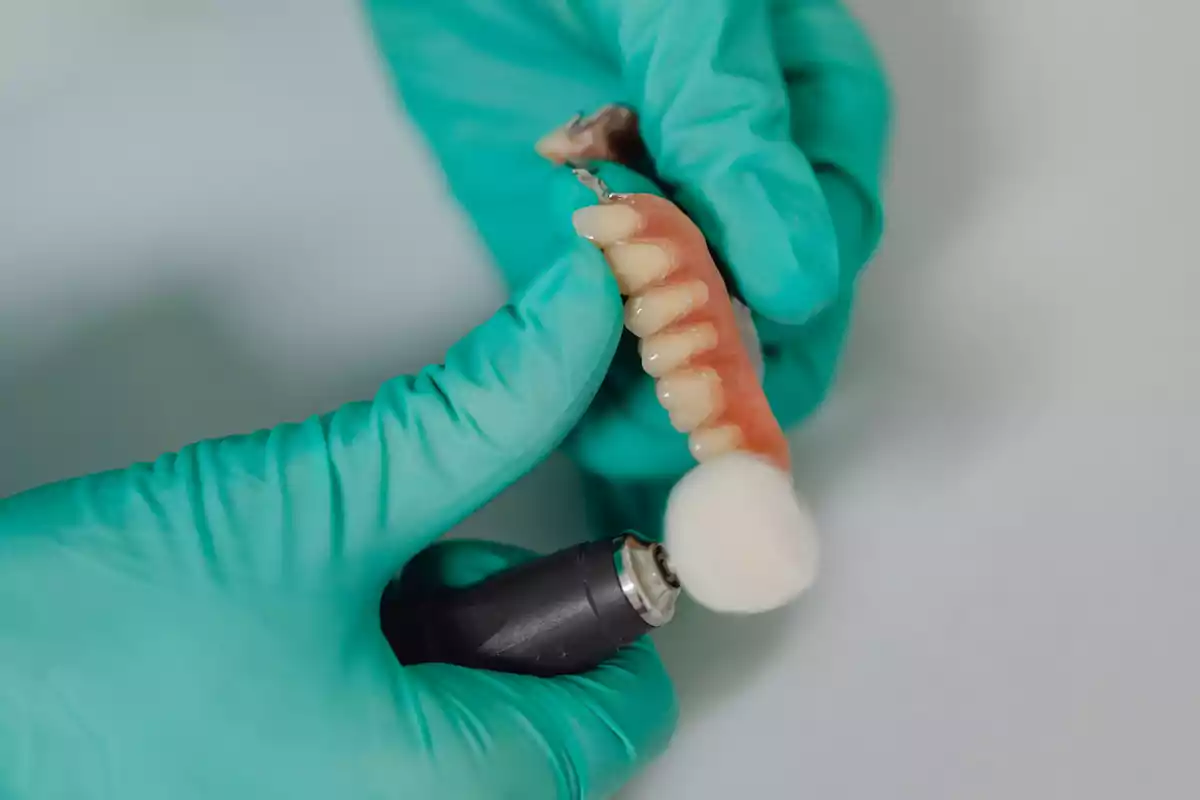 Persona con guantes verdes limpiando una dentadura postiza con una herramienta rotativa.