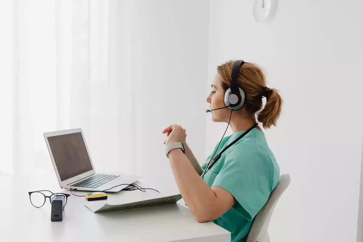 Una profesional de la salud con auriculares y micrófono trabaja en una videollamada frente a una computadora portátil en una oficina iluminada.