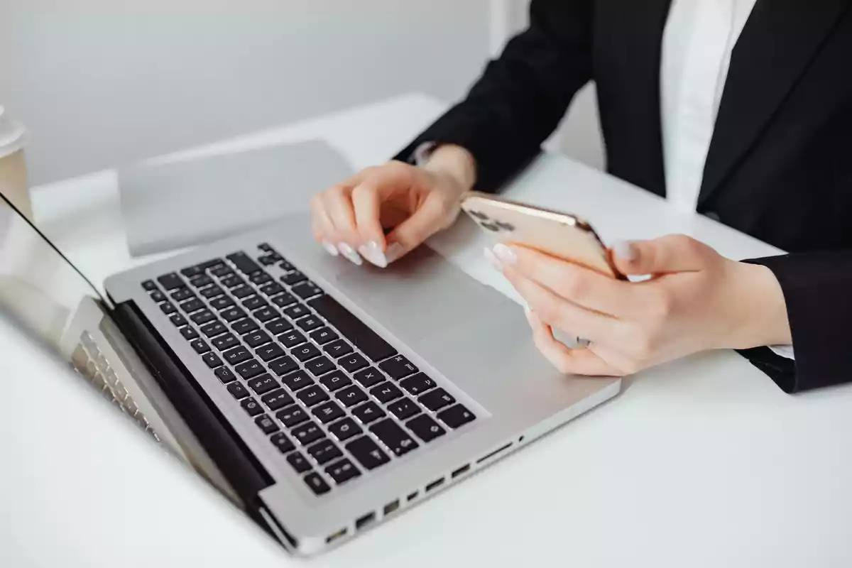 Persona usando un teléfono móvil y una computadora portátil en un escritorio blanco.