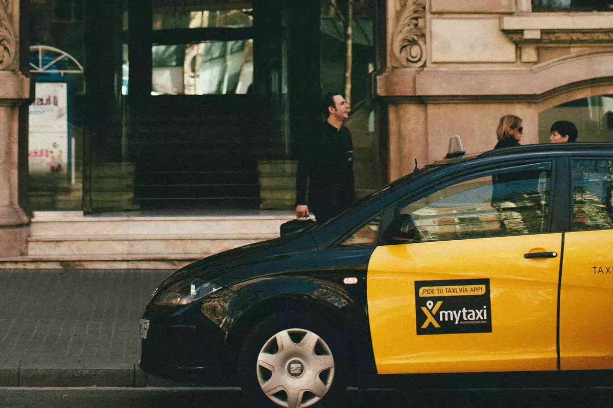 Taxi negro y amarillo estacionado en la calle frente a un edificio mientras un hombre camina con una maleta y dos mujeres conversan en la acera