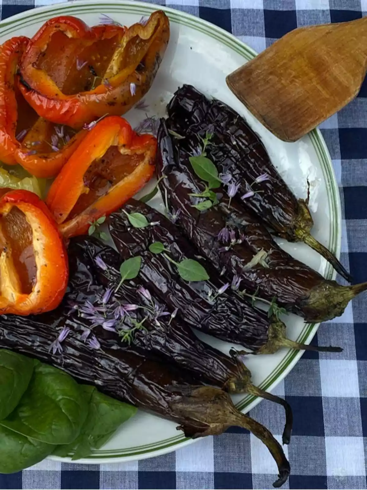 Un plato de pimientos rojos y berenjenas asadas adornado con hierbas frescas sobre un mantel de cuadros azules y blancos.