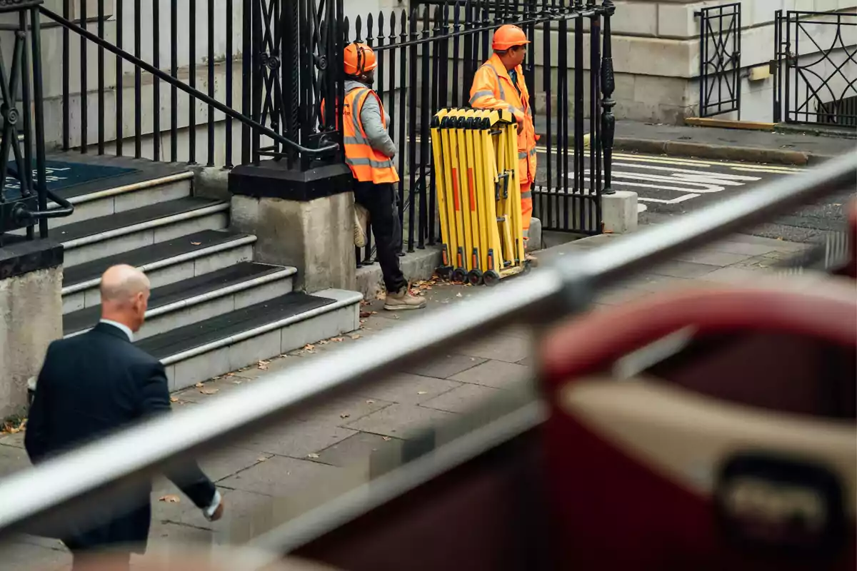 Un hombre de traje camina por la acera mientras dos trabajadores con chalecos y cascos naranjas están junto a una barrera amarilla frente a unas escaleras y una reja negra.