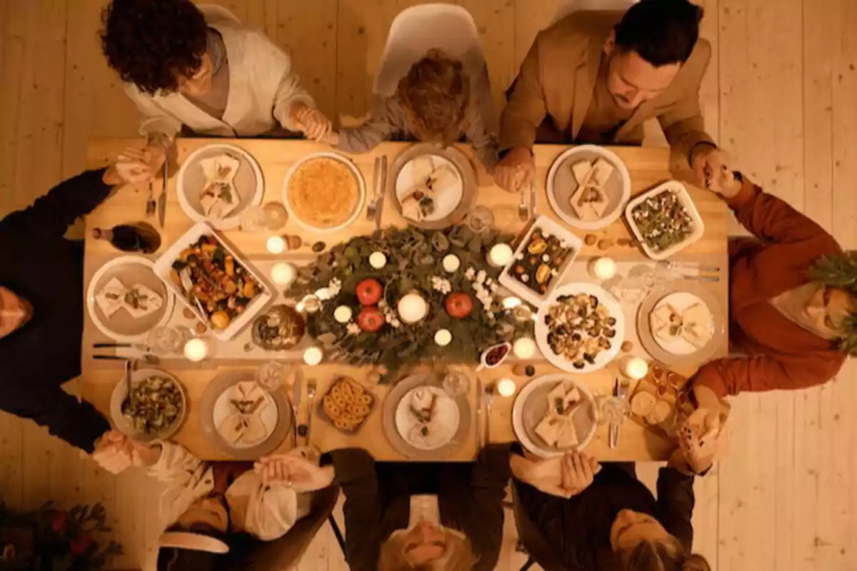 Personas sentadas alrededor de una mesa decorada con velas y comida, tomadas de las manos en un ambiente cálido y festivo.