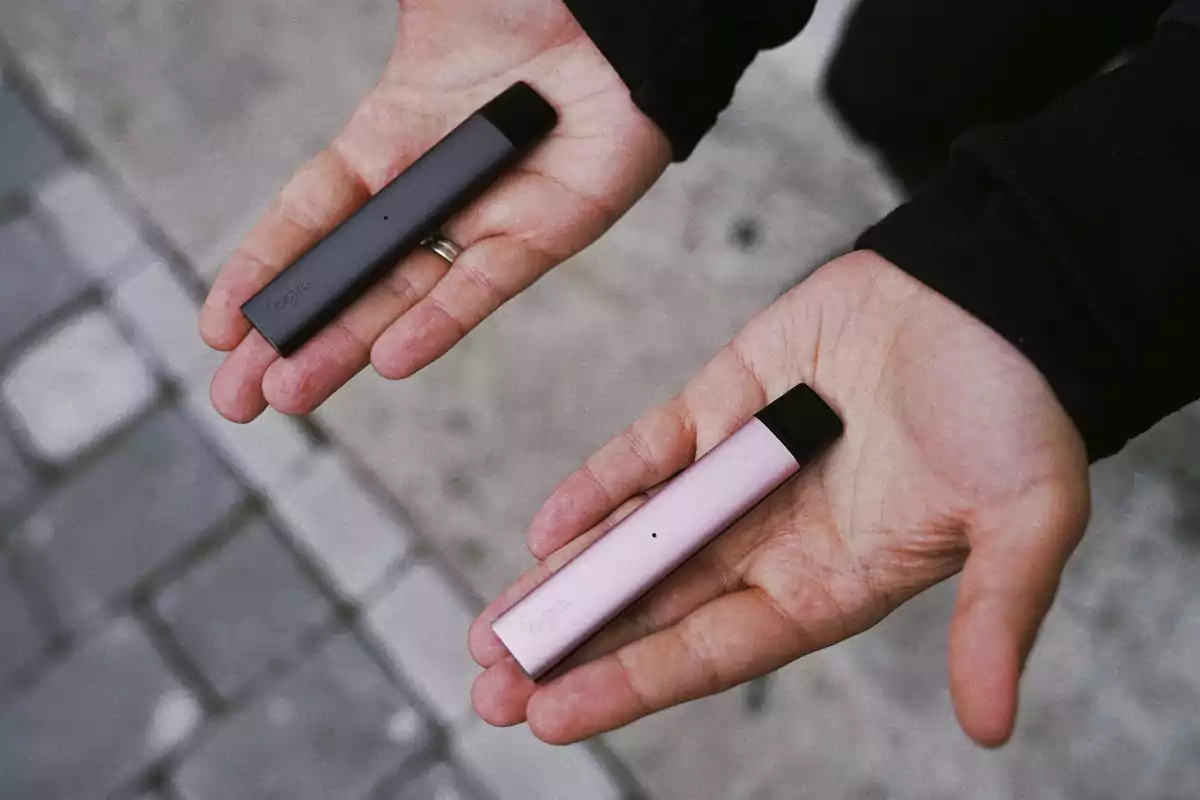 Dos manos sostienen cada una un vapeador de diferentes colores sobre un fondo de pavimento.
