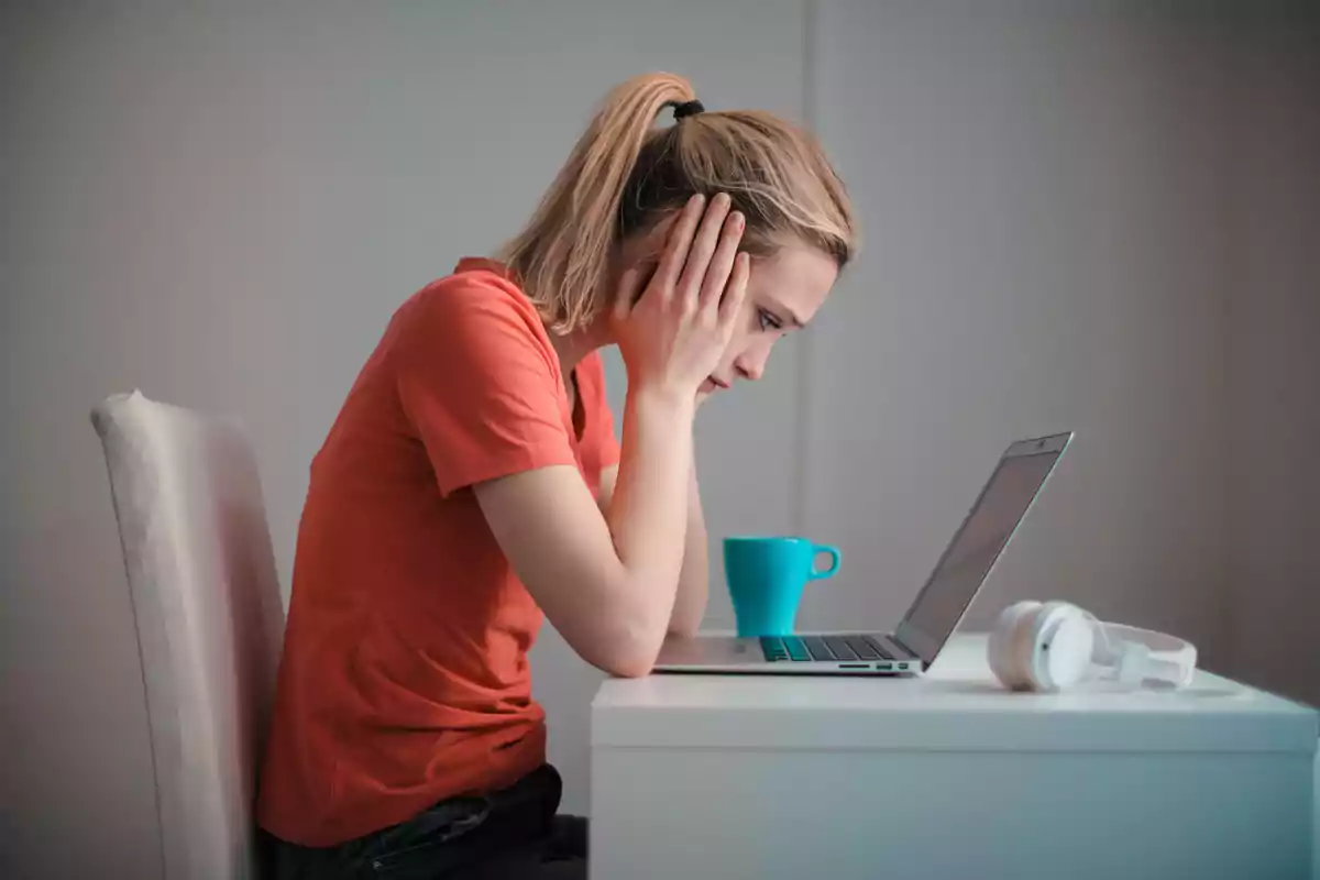 Persona con camiseta naranja sentada frente a una computadora portátil con una expresión de preocupación, con una taza azul y unos auriculares blancos sobre la mesa.