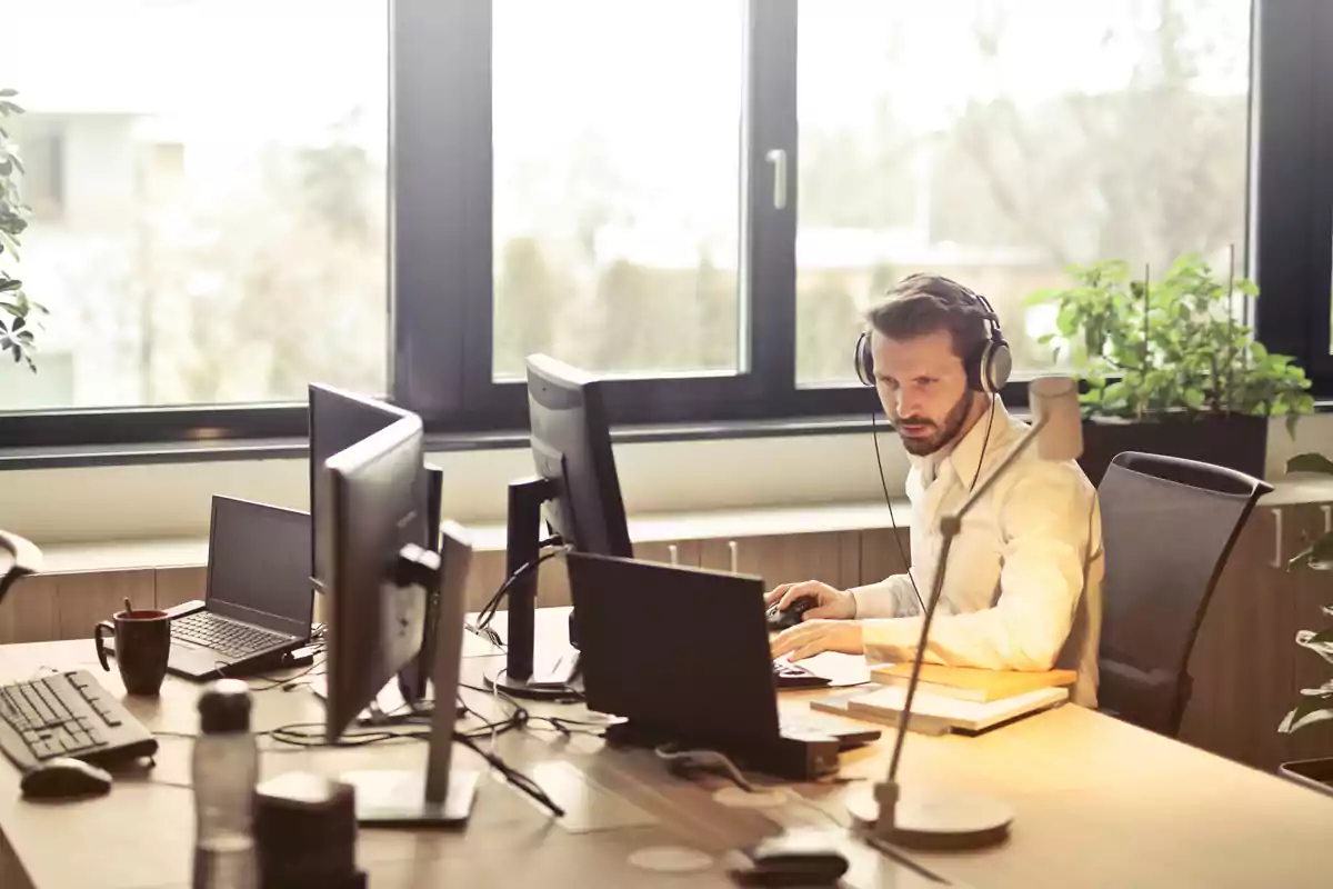 Hombre con auriculares trabajando en una oficina con varios monitores y una ventana grande al fondo.