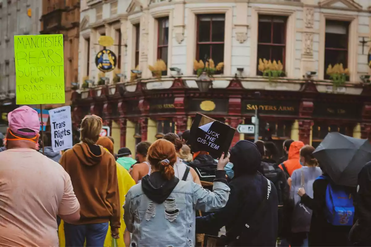 Un grupo de personas participa en una manifestación en la calle, sosteniendo carteles con mensajes de apoyo a los derechos trans frente a un edificio antiguo. Un grupo de personas participa en una manifestación en la calle, sosteniendo carteles con mensajes de apoyo a los derechos trans frente a un edificio antiguo.