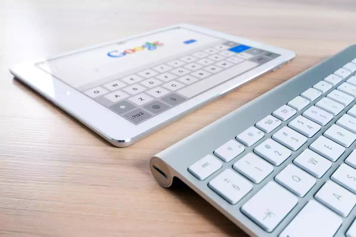 Una tableta con el buscador de Google en pantalla junto a un teclado inalámbrico sobre una mesa de madera.