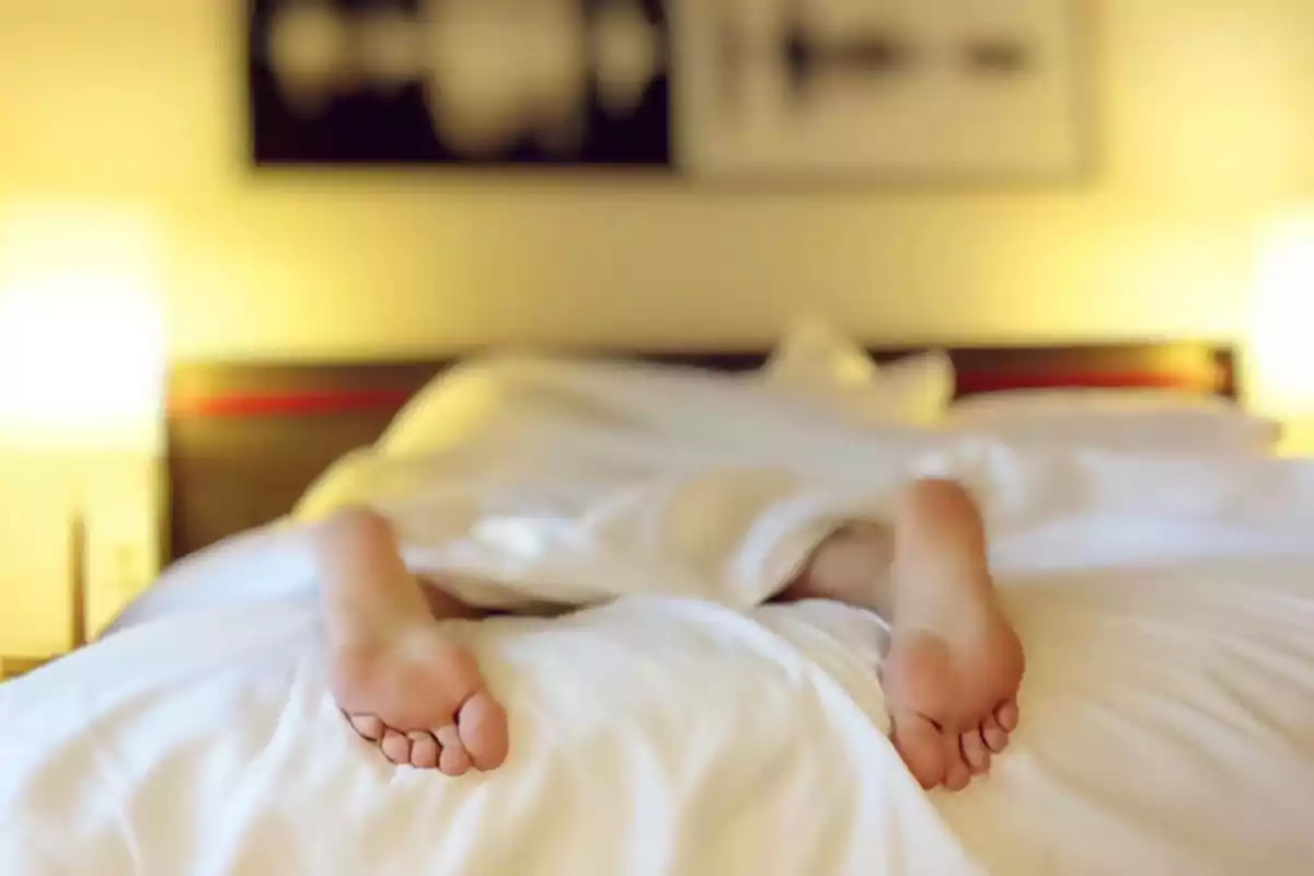 Persona acostada boca arriba en una cama con sábanas blancas y los pies descubiertos