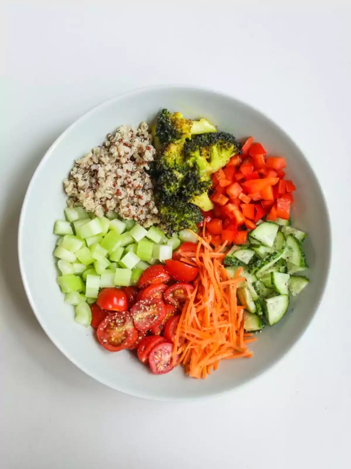 Un plato de ensalada con quinoa, brócoli, pimientos rojos, pepino, zanahoria rallada, tomates cherry y apio.