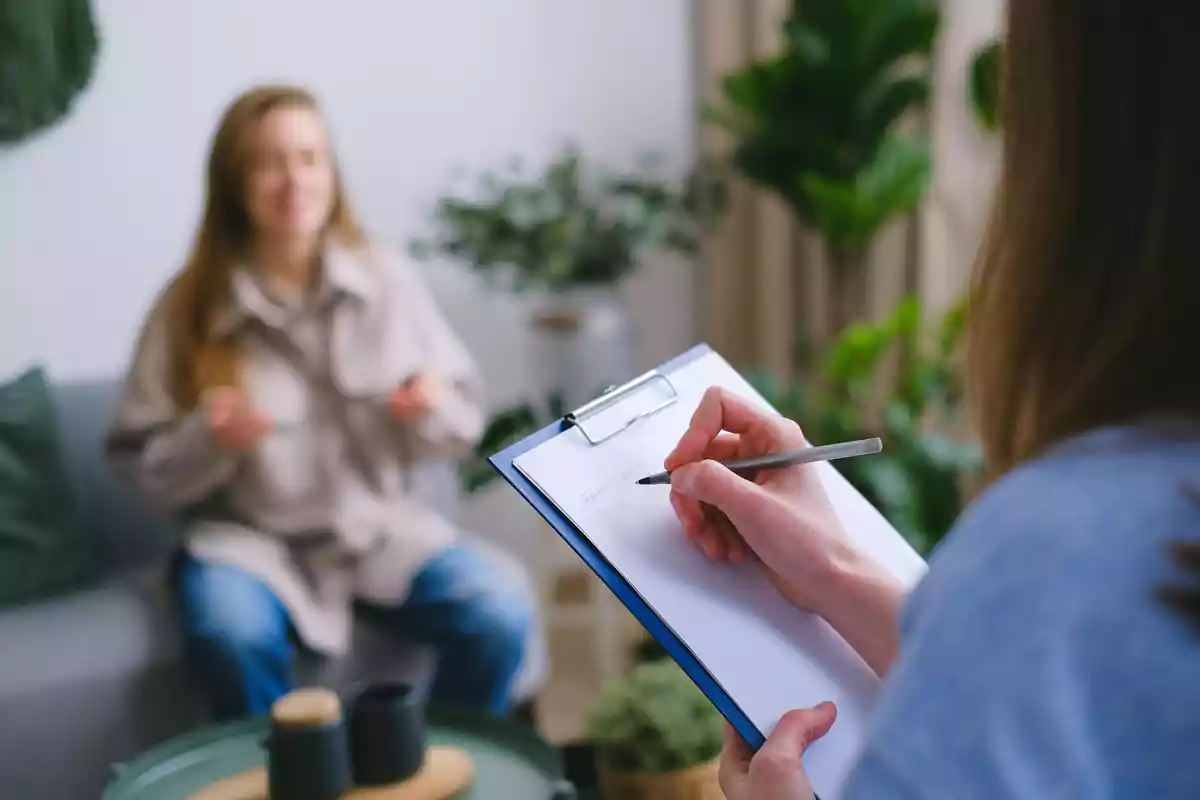 Una persona tomando notas en un portapapeles mientras otra persona habla en un entorno acogedor con plantas.