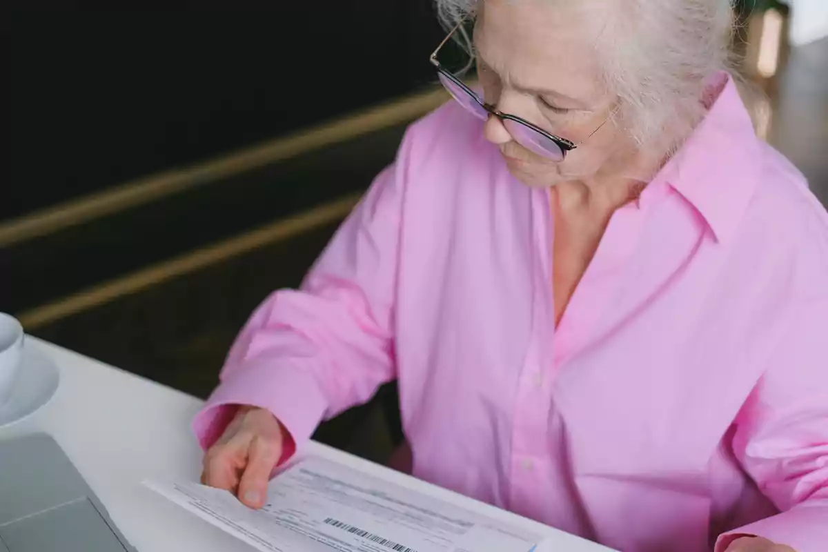 Mujer mayor con camisa rosa revisando documentos en una mesa.