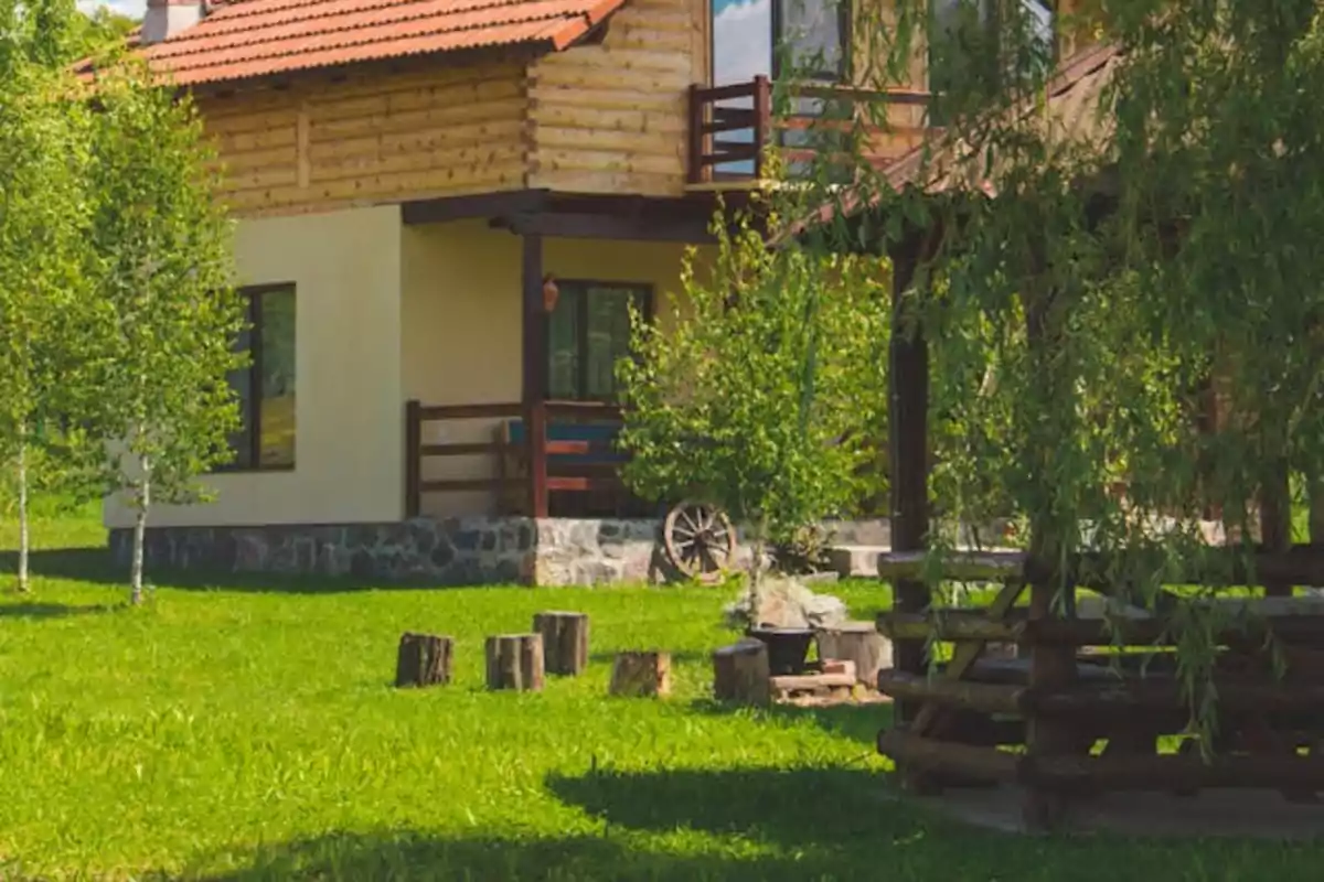 Casa de campo con techo de tejas rojas, paredes de madera y piedra, rodeada de árboles y césped verde, con un porche y una estructura de madera en el jardín.