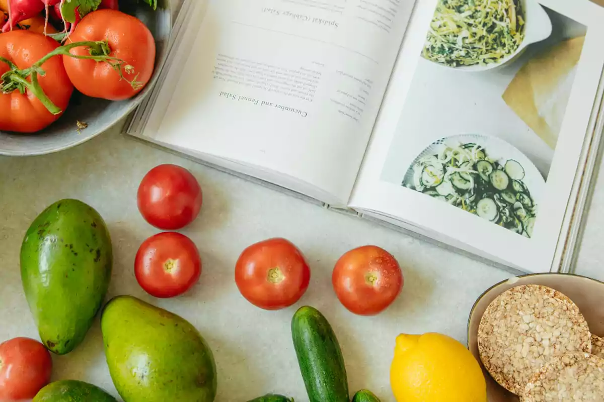 Un libro de cocina abierto junto a tomates, aguacates, pepinos, un limón y galletas de arroz sobre una mesa.