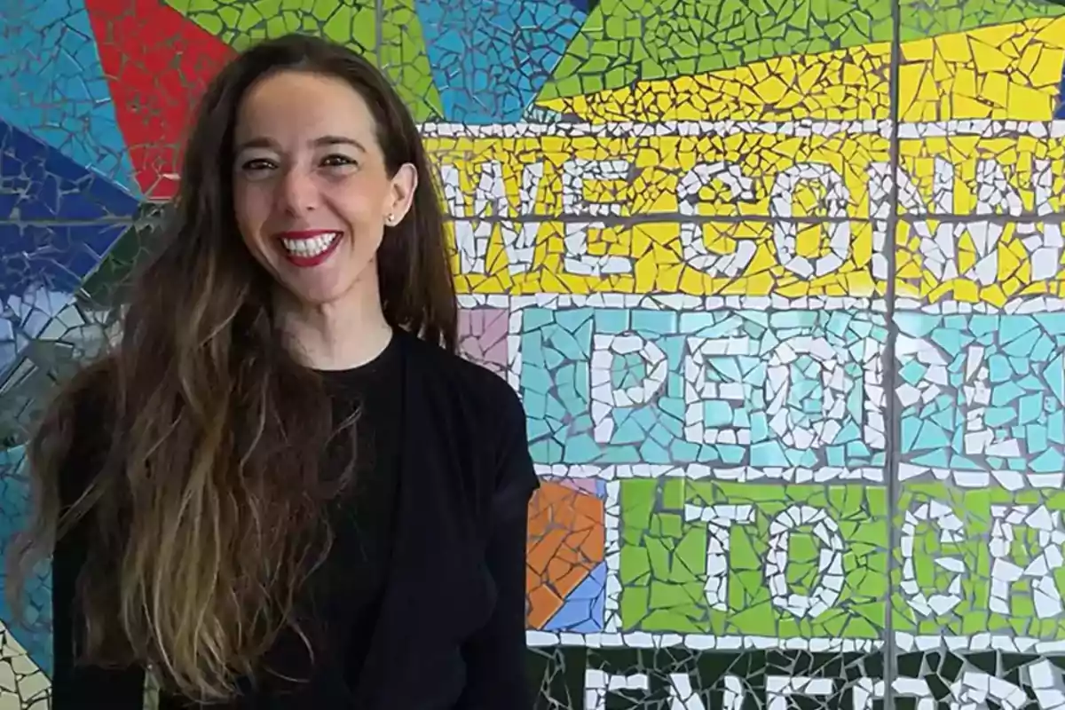 Una mujer sonriente con cabello largo y suelto está de pie frente a un colorido mural de mosaico que tiene texto en inglés.