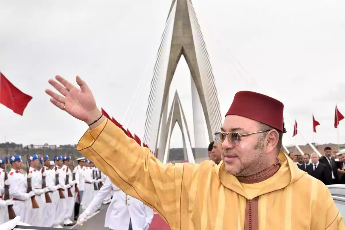 Un hombre con gafas de sol y un fez rojo saluda mientras camina por un puente moderno, acompañado por una guardia de honor vestida de blanco y banderas rojas ondeando en el fondo.