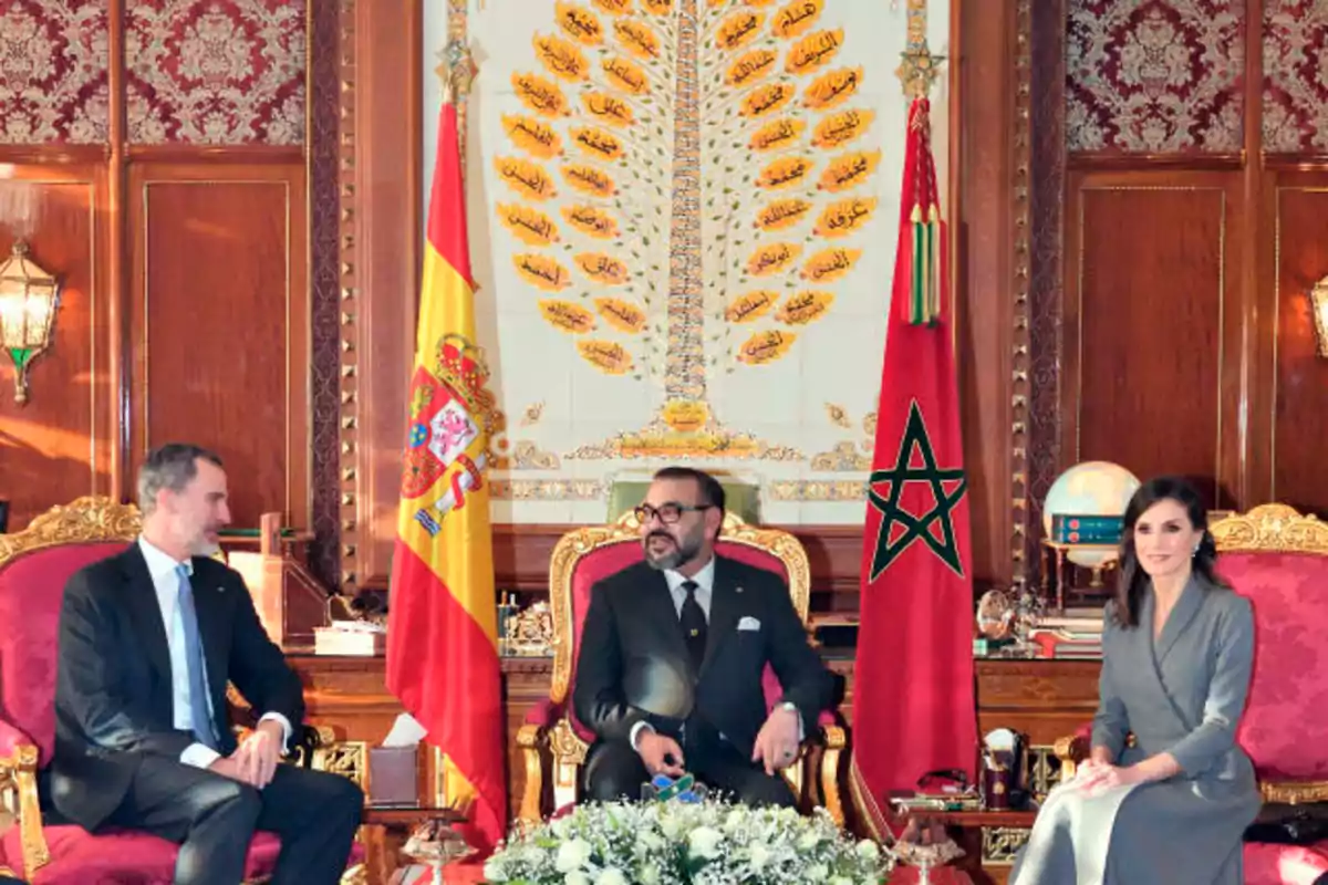 Tres personas sentadas en sillas doradas y rojas, con las banderas de España y Marruecos detrás de ellos, en una sala decorada con motivos árabes.