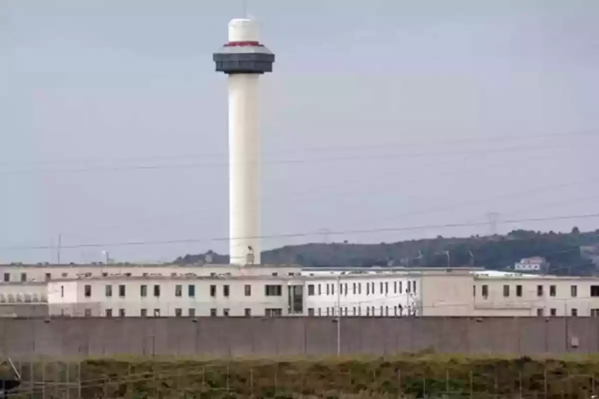 Una torre de vigilancia se eleva sobre un edificio rodeado de un muro alto en un entorno rural.