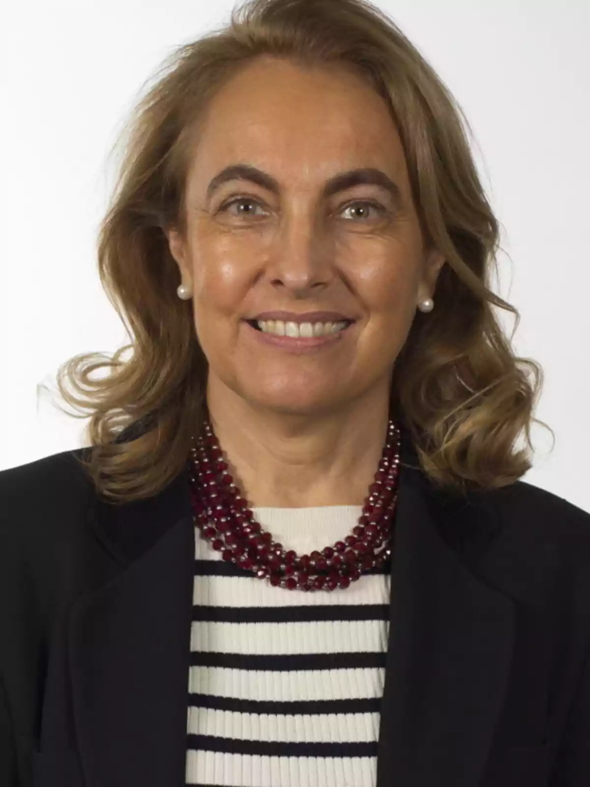 Una persona sonriendo con cabello castaño claro y rizado, vistiendo un collar de perlas rojas, un suéter de rayas blancas y negras, y un saco oscuro. Una persona sonriendo con cabello castaño claro y rizado, vistiendo un collar de perlas rojas, un suéter de rayas blancas y negras, y un saco oscuro.
