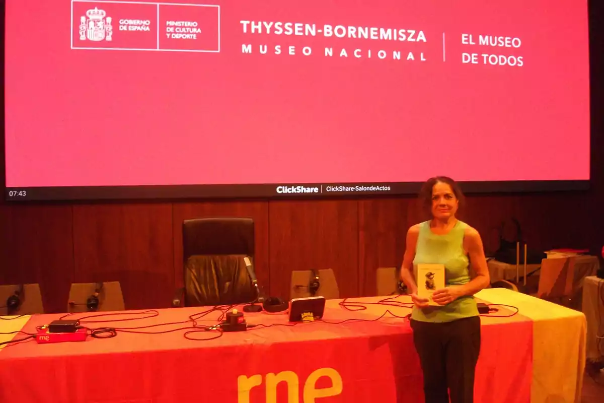 Mujer de pie sosteniendo un libro frente a una mesa con equipo de audio en un auditorio con una pantalla grande de fondo que muestra el nombre del Museo Nacional Thyssen-Bornemisza y el lema El museo de todos