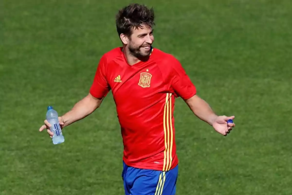 Un jugador de fútbol con camiseta roja y pantalones cortos azules sostiene una botella de agua en un campo de césped.