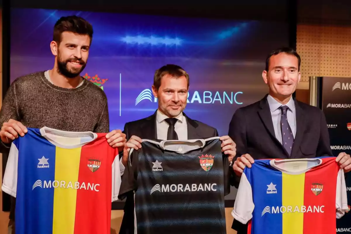 Tres hombres sostienen camisetas de fútbol con el logo de Morabanc en un evento.