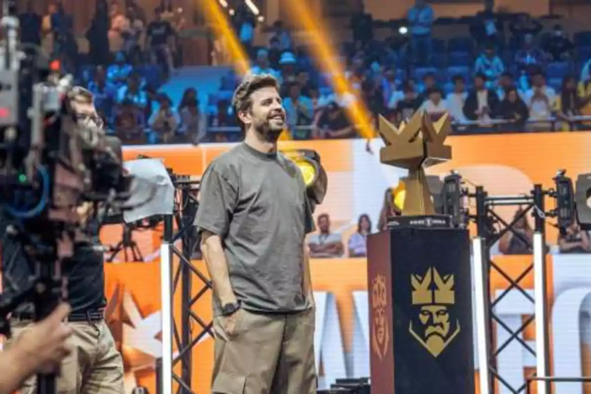 Un hombre sonriente de pie en un escenario junto a un trofeo dorado con forma de corona mientras una multitud observa en el fondo