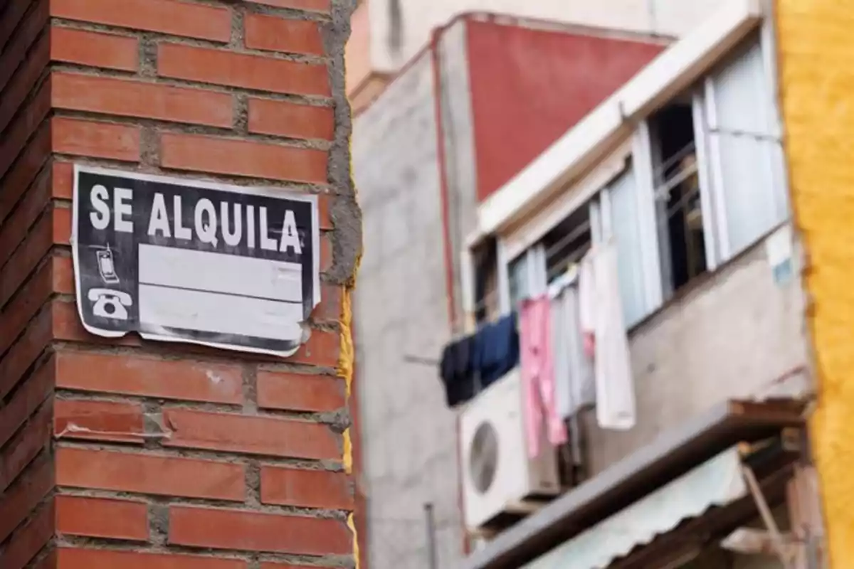 Un cartel de "Se Alquila" en una pared de ladrillo con un edificio al fondo.