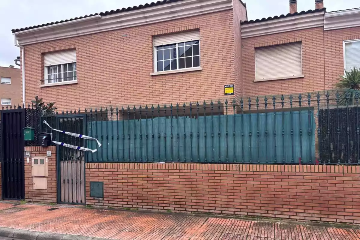 Una casa de ladrillo con ventanas cerradas y una cerca de metal verde al frente, con cinta de advertencia en la puerta.