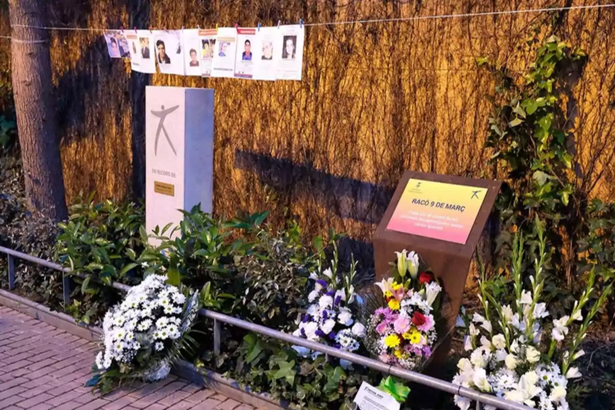 Un monumento conmemorativo rodeado de flores y fotografías colgadas en una cuerda, ubicado en un entorno natural con vegetación.