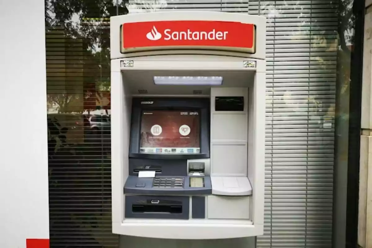 Cajero automático del banco Santander en una calle con persianas metálicas de fondo.