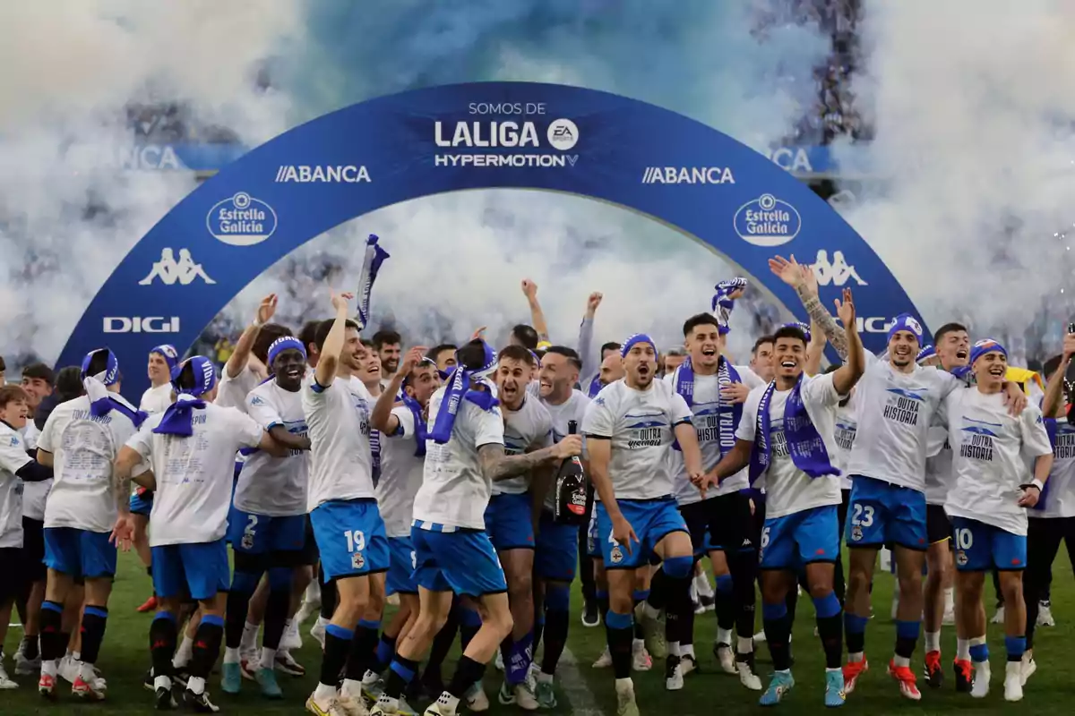 Jugadores de fútbol celebrando bajo un arco azul con humo en el fondo.