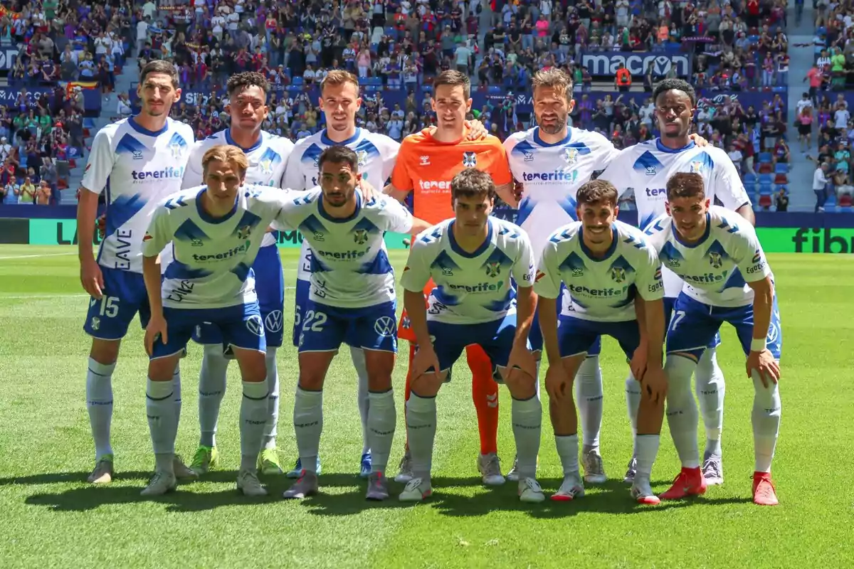 Un equipo de fútbol posando en el campo antes de un partido, con jugadores vistiendo uniformes blancos y azules y un portero con uniforme naranja, mientras el público observa desde las gradas. Un equipo de fútbol posando en el campo antes de un partido, con jugadores vistiendo uniformes blancos y azules y un portero con uniforme naranja, mientras el público observa desde las gradas.