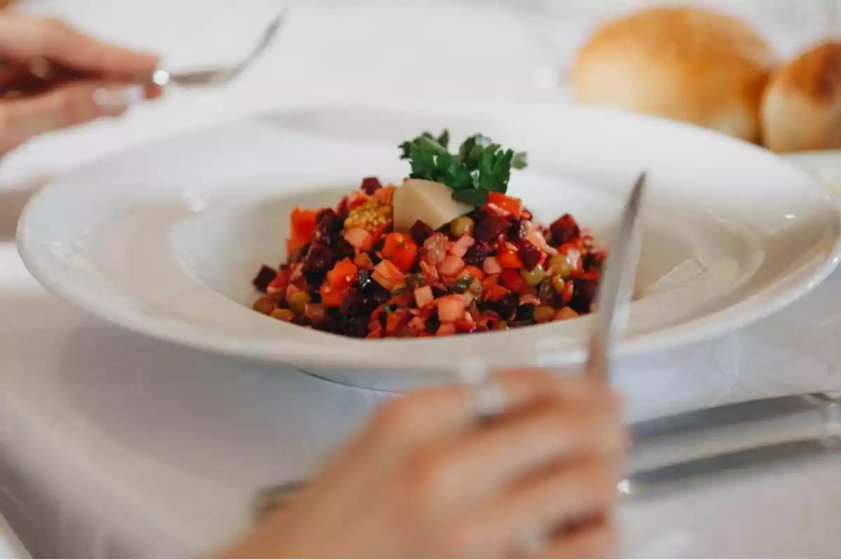 Un plato de ensalada colorida con vegetales picados y una guarnición de perejil en el centro, servido en un restaurante con panecillos en el fondo y una persona sosteniendo cubiertos.