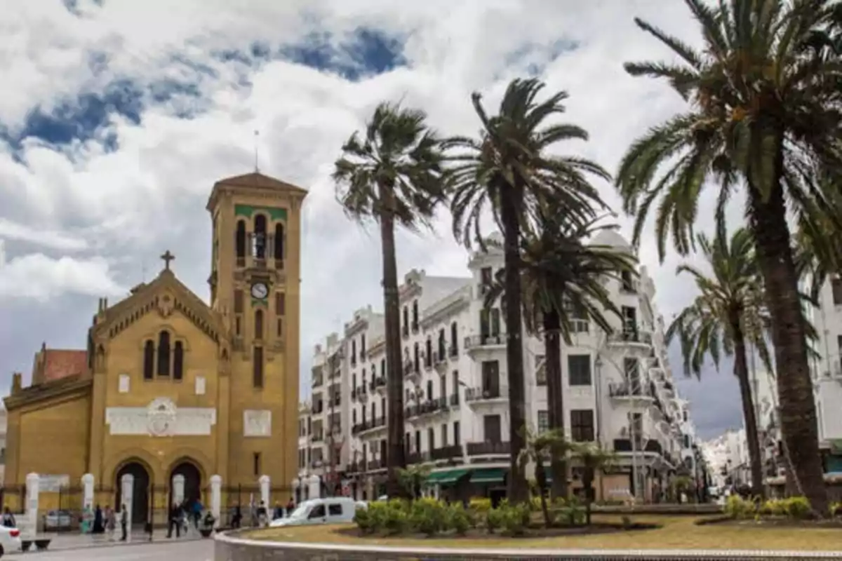 Una iglesia con una torre de reloj se encuentra en una plaza rodeada de palmeras y edificios blancos bajo un cielo parcialmente nublado.