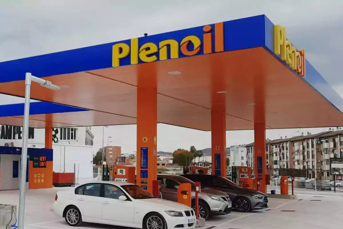 Estación de servicio Plenoil con varios coches repostando bajo un techo naranja y azul en una zona urbana Estación de servicio Plenoil con varios coches repostando bajo un techo naranja y azul en una zona urbana