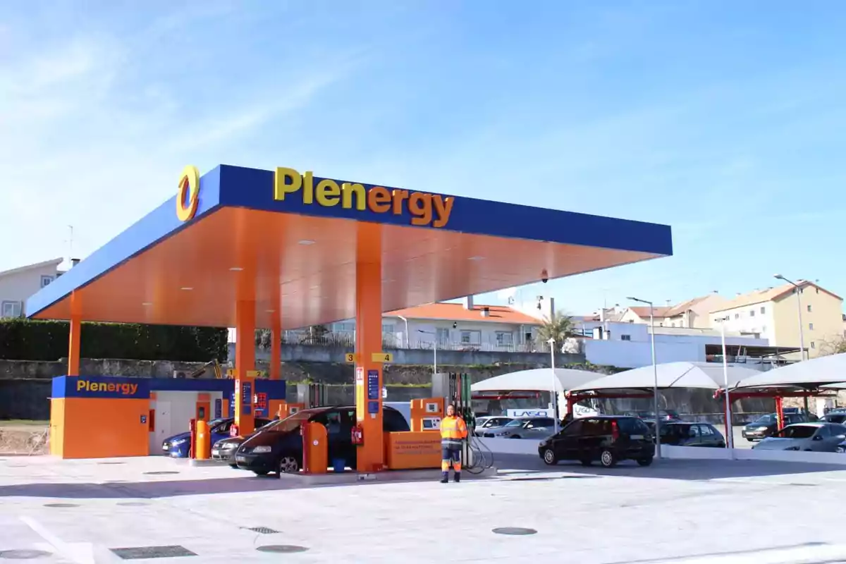 Estación de servicio Plenergy con techo azul y naranja, varios autos repostando y un trabajador con chaleco reflectante Estación de servicio Plenergy con techo azul y naranja, varios autos repostando y un trabajador con chaleco reflectante