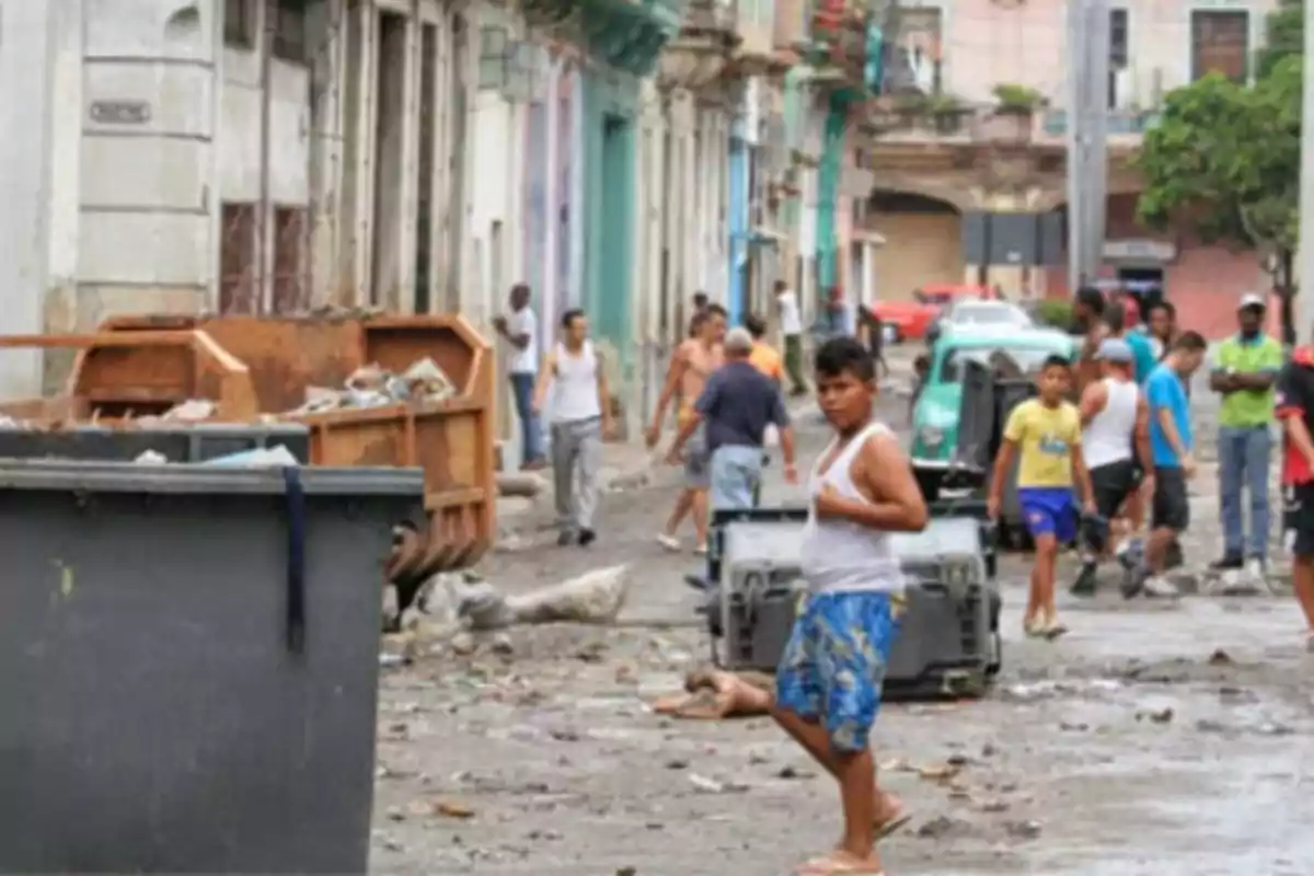 Personas caminando por una calle con basura y contenedores en un entorno urbano deteriorado.